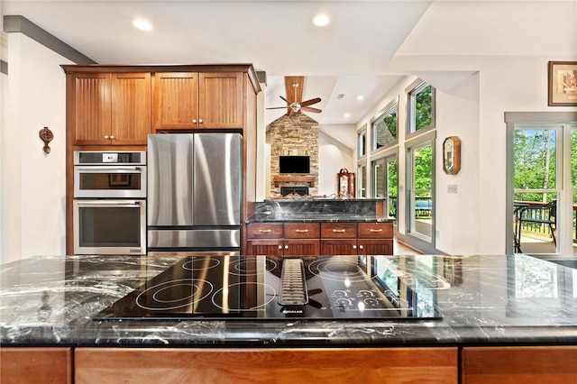 kitchen featuring brown cabinetry, a spacious island, ceiling fan, a stone fireplace, and appliances with stainless steel finishes
