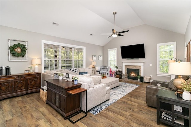 living area with a ceiling fan, lofted ceiling, a healthy amount of sunlight, a glass covered fireplace, and wood finished floors