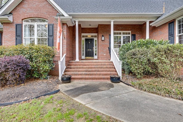 entrance to property featuring a shingled roof, brick siding, and covered porch