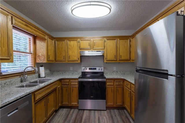 kitchen with stainless steel appliances, a sink, under cabinet range hood, brown cabinetry, and dark wood finished floors