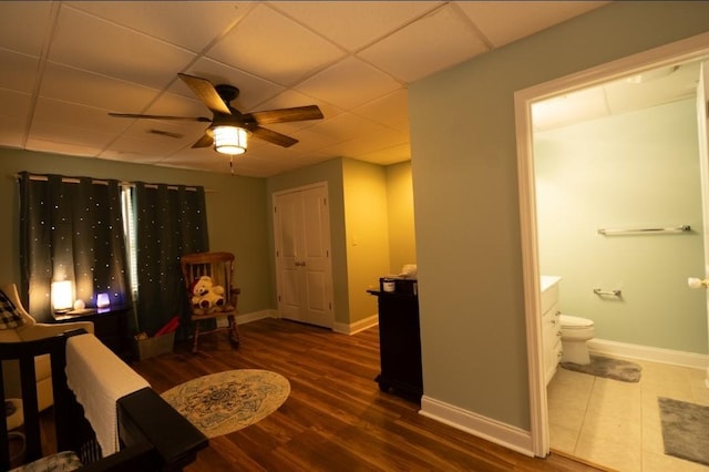 bedroom featuring baseboards, a drop ceiling, ensuite bathroom, wood finished floors, and a ceiling fan