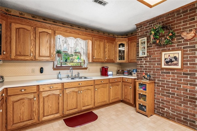 kitchen with a sink, brown cabinets, light countertops, brick wall, and glass insert cabinets