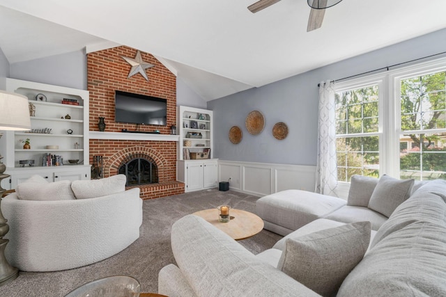 carpeted living room featuring a fireplace, vaulted ceiling, ceiling fan, and wainscoting