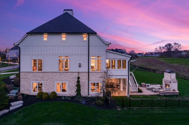 back of house at dusk with a patio area, fence private yard, a lit fireplace, and a lawn