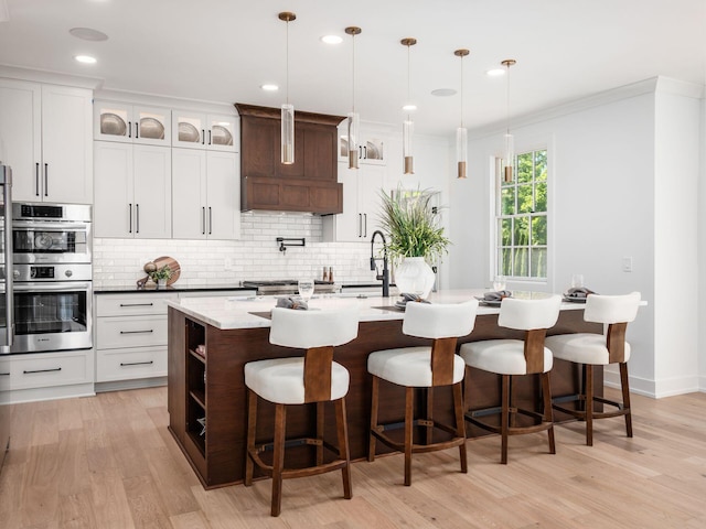 kitchen with hanging light fixtures, stainless steel double oven, light hardwood / wood-style floors, and a kitchen island with sink