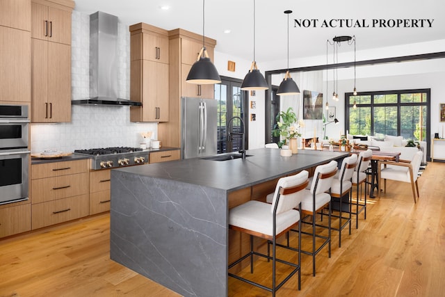 kitchen with a center island with sink, dark countertops, decorative light fixtures, wall chimney range hood, and a sink