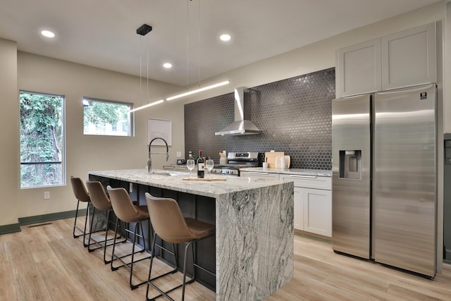 kitchen with wall chimney exhaust hood, stainless steel appliances, a kitchen island with sink, light hardwood / wood-style flooring, and white cabinetry