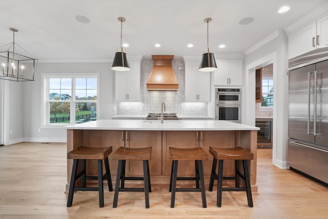 kitchen with custom exhaust hood, a kitchen island with sink, white cabinets, light hardwood / wood-style flooring, and stainless steel appliances