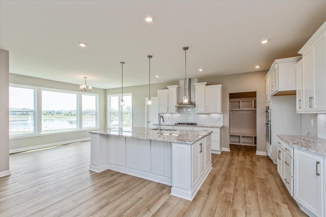 kitchen featuring sink, wall chimney exhaust hood, an island with sink, pendant lighting, and white cabinets