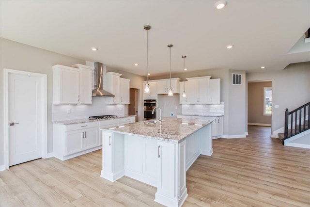 kitchen featuring light stone countertops, appliances with stainless steel finishes, wall chimney exhaust hood, white cabinetry, and an island with sink