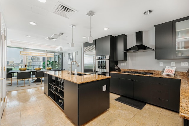 kitchen with gas cooktop, sink, wall chimney range hood, pendant lighting, and a center island with sink