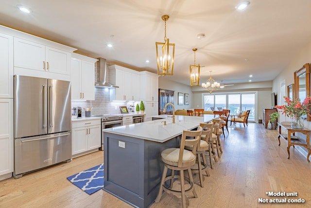 kitchen featuring stainless steel appliances, wall chimney range hood, a center island with sink, white cabinets, and hanging light fixtures