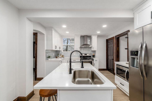 kitchen featuring a kitchen island with sink, sink, wall chimney exhaust hood, appliances with stainless steel finishes, and white cabinetry