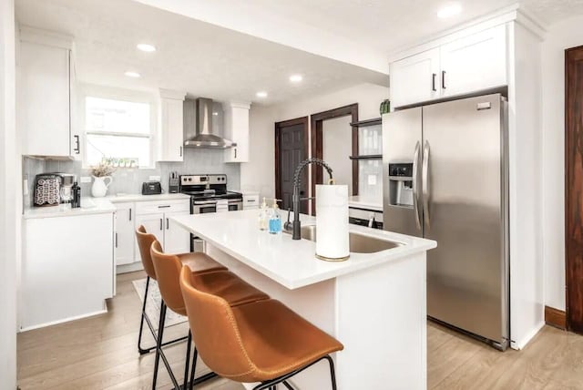 kitchen with a kitchen island with sink, wall chimney exhaust hood, light wood-type flooring, a kitchen bar, and stainless steel appliances