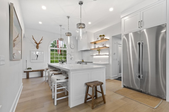 kitchen with hanging light fixtures, light hardwood / wood-style floors, a kitchen island with sink, stainless steel built in fridge, and white cabinets