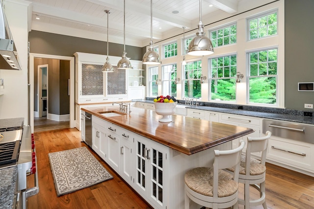 kitchen with wooden counters, a center island with sink, wood-type flooring, beamed ceiling, and white cabinetry