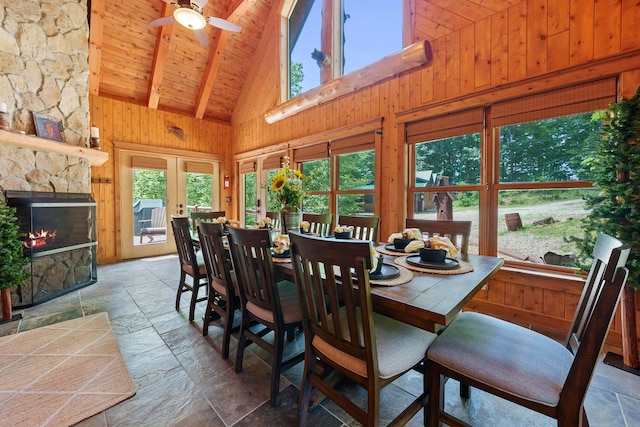 dining space featuring a stone fireplace, beamed ceiling, high vaulted ceiling, wooden walls, and wood ceiling