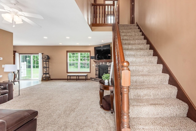 staircase featuring a stone fireplace, ceiling fan, plenty of natural light, and carpet