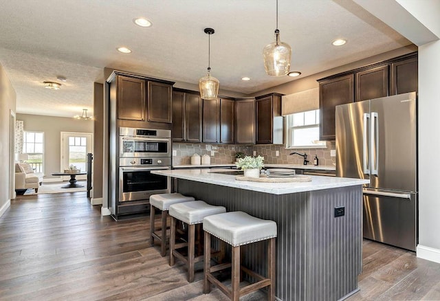 kitchen with dark wood-style flooring, dark brown cabinets, appliances with stainless steel finishes, a center island, and tasteful backsplash