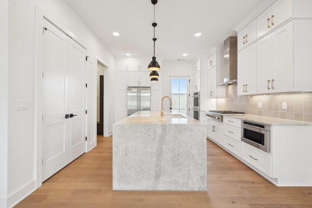 kitchen with wall chimney range hood, a sink, built in appliances, backsplash, and white cabinetry