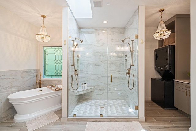 bathroom with a skylight, an inviting chandelier, stacked washer / dryer, tile walls, and shower with separate bathtub