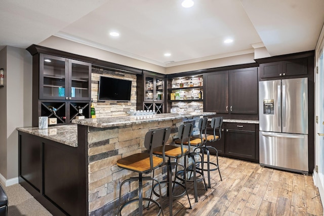 bar featuring crown molding, stainless steel refrigerator with ice dispenser, light stone countertops, light wood-type flooring, and dark brown cabinetry