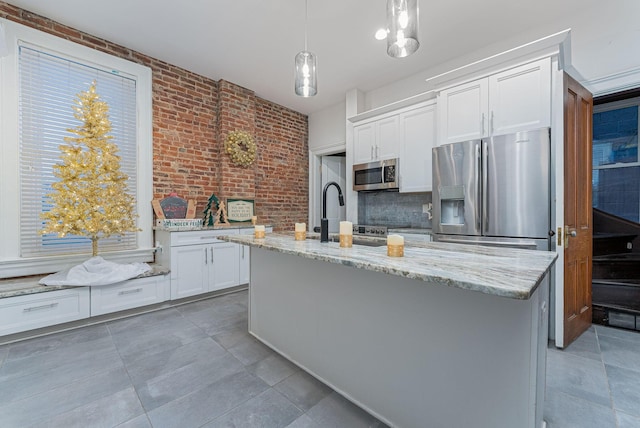 kitchen featuring a center island with sink, appliances with stainless steel finishes, light stone counters, white cabinetry, and brick wall