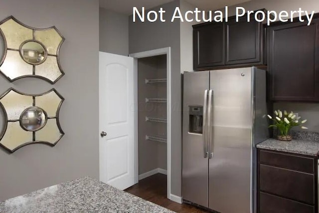 kitchen with stainless steel fridge, dark brown cabinetry, light stone counters, and dark wood-type flooring