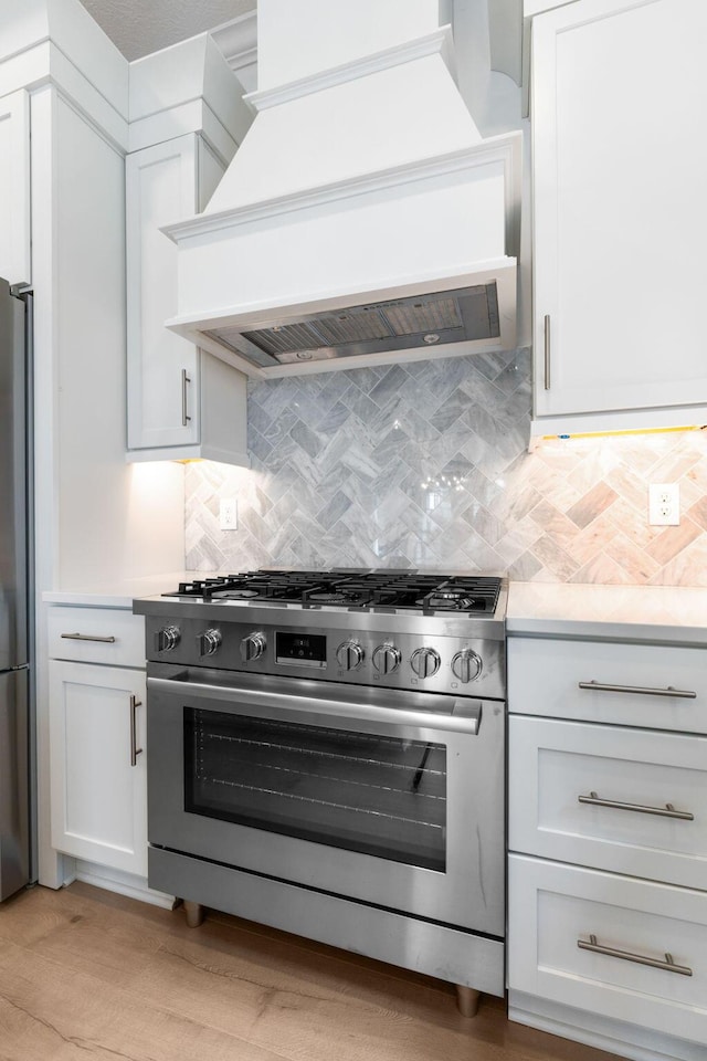 kitchen with custom exhaust hood, white cabinets, light hardwood / wood-style flooring, decorative backsplash, and appliances with stainless steel finishes