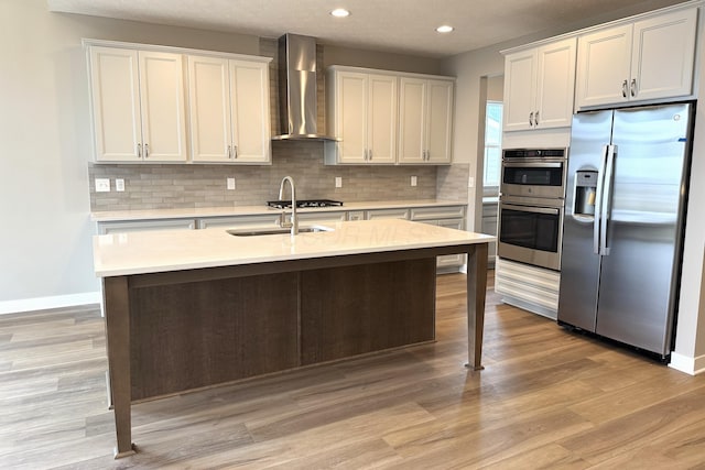 kitchen featuring wall chimney range hood, sink, appliances with stainless steel finishes, white cabinetry, and an island with sink