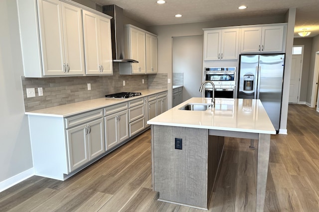 kitchen with appliances with stainless steel finishes, sink, hardwood / wood-style flooring, a center island with sink, and wall chimney range hood