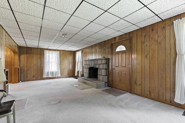 unfurnished living room with light colored carpet, a stone fireplace, a drop ceiling, and wooden walls