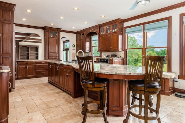 kitchen with light stone countertops, backsplash, a center island with sink, and crown molding