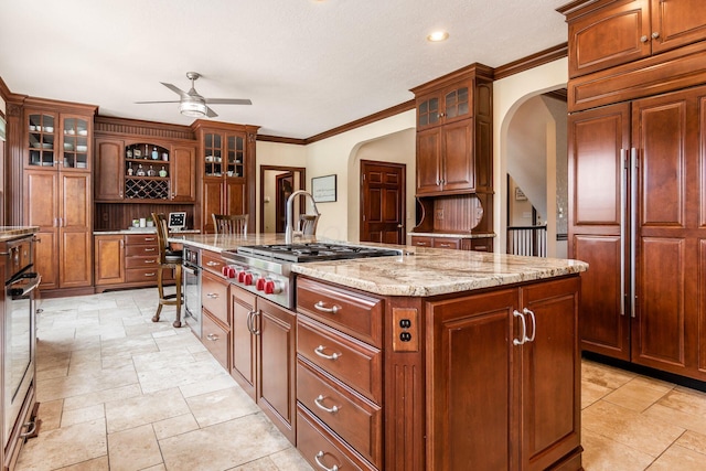 kitchen with light stone countertops, ceiling fan, stainless steel appliances, a center island with sink, and ornamental molding