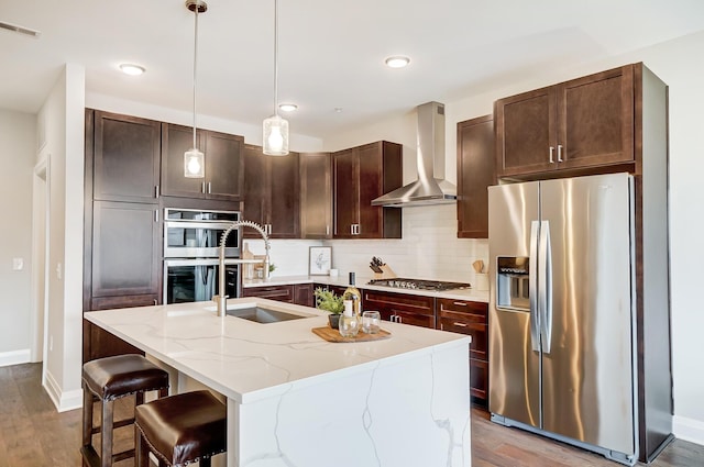 kitchen with a kitchen island with sink, hanging light fixtures, hardwood / wood-style flooring, wall chimney exhaust hood, and appliances with stainless steel finishes