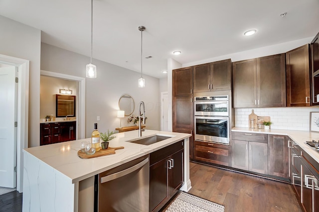 kitchen with dark wood-type flooring, a center island with sink, hanging light fixtures, sink, and appliances with stainless steel finishes