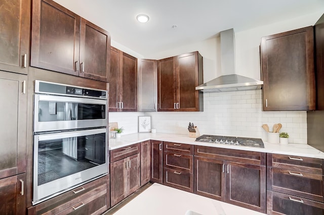 kitchen with dark brown cabinetry, wall chimney range hood, stainless steel appliances, and tasteful backsplash