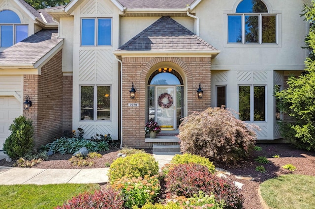 view of exterior entry with a garage, brick siding, roof with shingles, and stucco siding