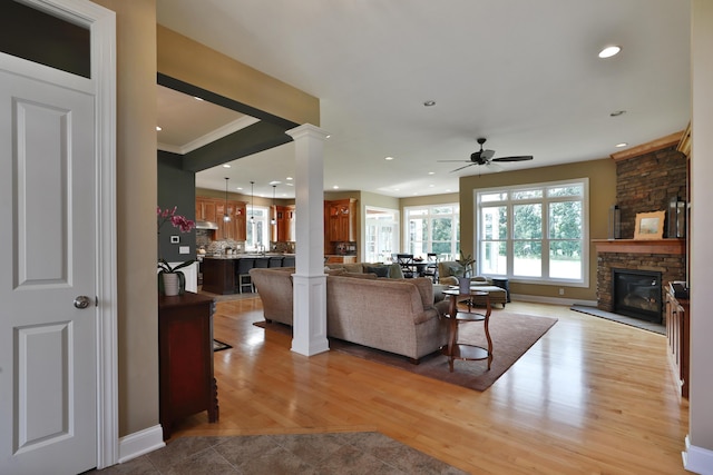 living room featuring ceiling fan, a stone fireplace, light hardwood / wood-style flooring, decorative columns, and ornamental molding