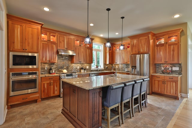 kitchen with light stone countertops, a breakfast bar, stainless steel appliances, hanging light fixtures, and an island with sink