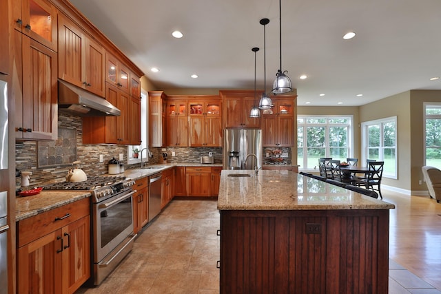 kitchen featuring stainless steel appliances, light stone counters, pendant lighting, decorative backsplash, and a center island with sink