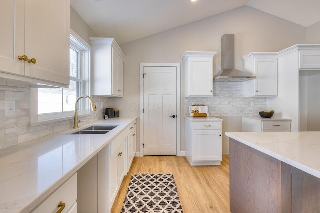 kitchen featuring light stone counters, a sink, white cabinets, vaulted ceiling, and wall chimney range hood