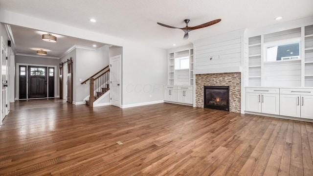 unfurnished living room with built in shelves, ceiling fan, dark wood-type flooring, a barn door, and a fireplace