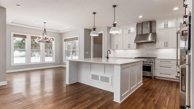 kitchen featuring wall chimney exhaust hood, high end stainless steel range oven, a kitchen island with sink, pendant lighting, and white cabinetry