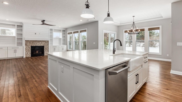 kitchen with a stone fireplace, stainless steel dishwasher, decorative light fixtures, a kitchen island with sink, and ceiling fan with notable chandelier