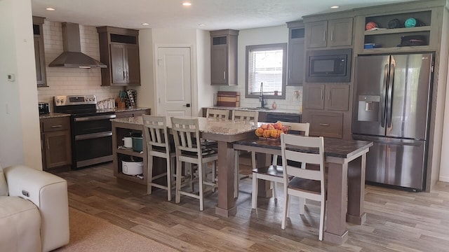 kitchen with wall chimney exhaust hood, tasteful backsplash, appliances with stainless steel finishes, a kitchen island, and light wood-type flooring