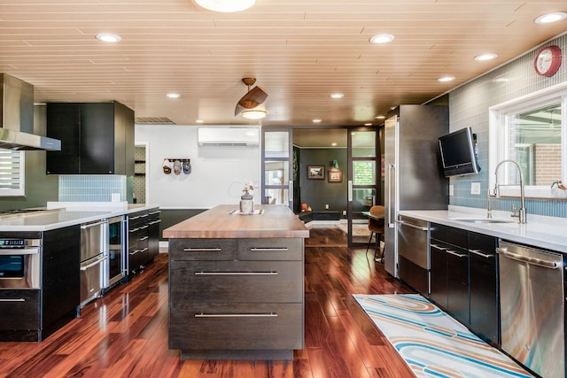 kitchen with a wall mounted air conditioner, butcher block countertops, a sink, stainless steel appliances, and wall chimney exhaust hood