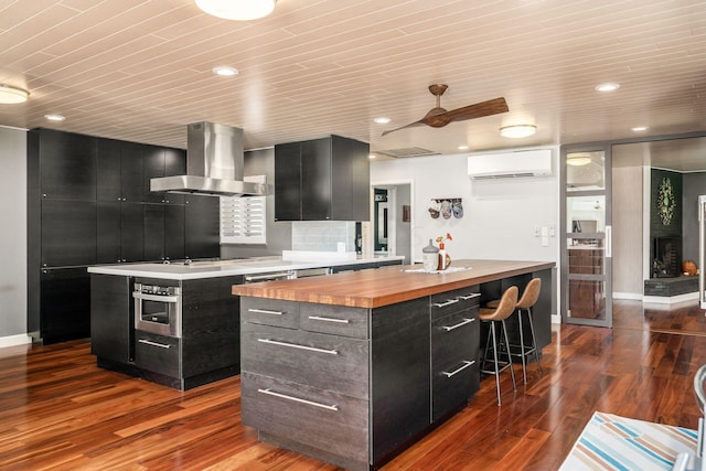 kitchen with dark cabinetry, a kitchen island, butcher block countertops, a wall mounted air conditioner, and wall chimney exhaust hood