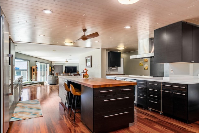 kitchen with dark cabinetry, a breakfast bar, dark wood-type flooring, wall chimney range hood, and butcher block counters