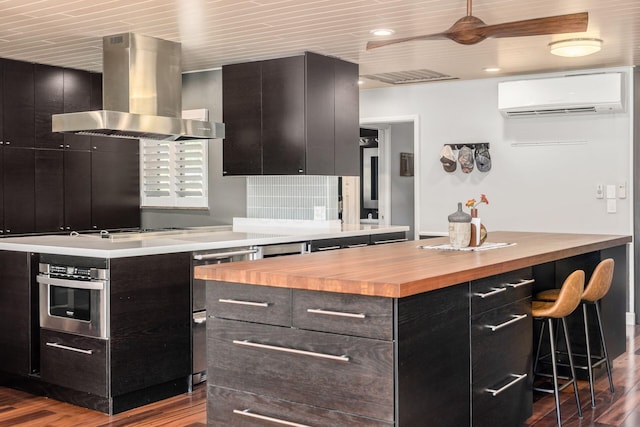 kitchen with an AC wall unit, stainless steel oven, wall chimney exhaust hood, wood counters, and dark cabinets